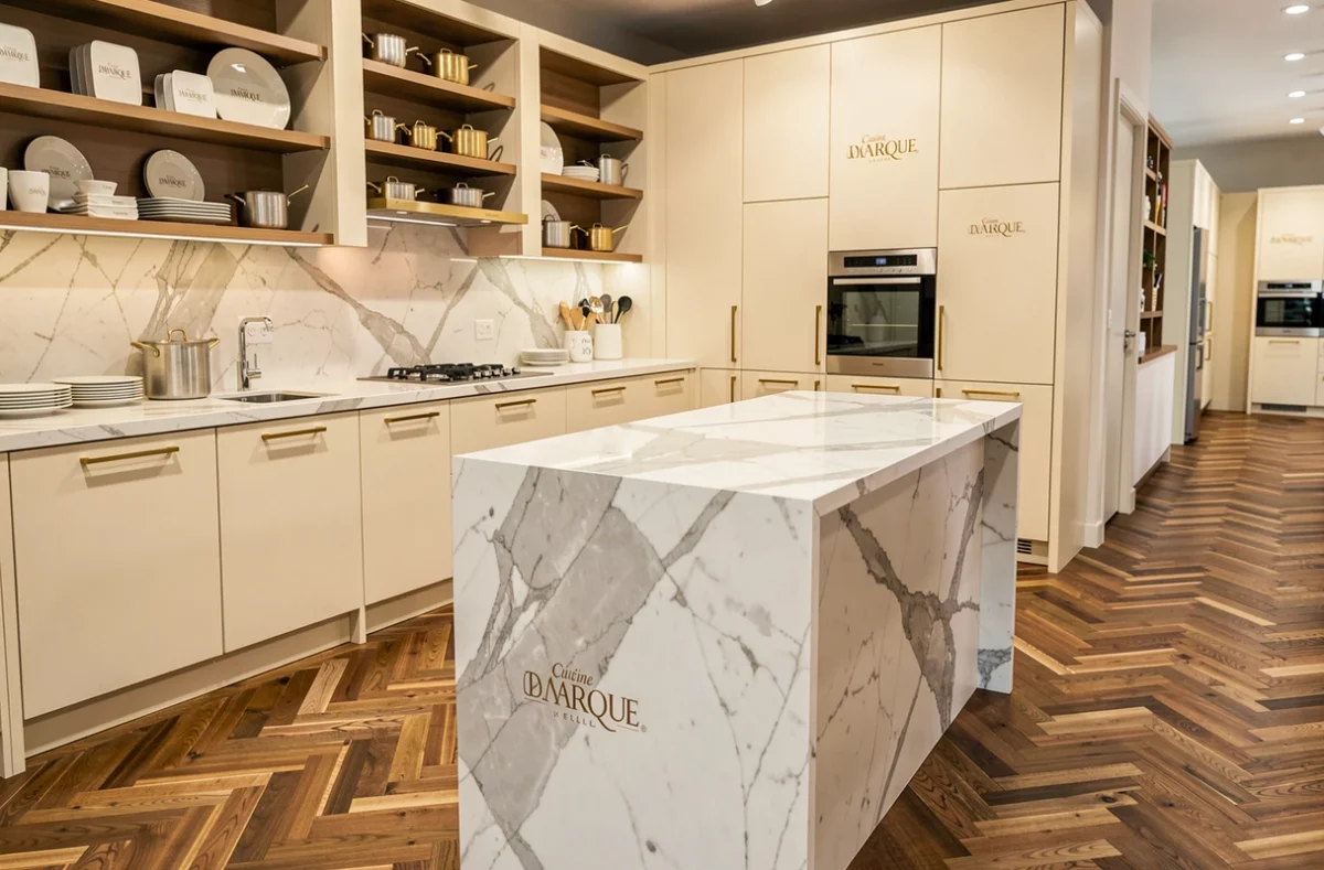 Showroom Cuisine de Marque Lille avec cuisine en laque crème, quincaillerie dorée brossée et îlot en marbre sur parquet en chevrons